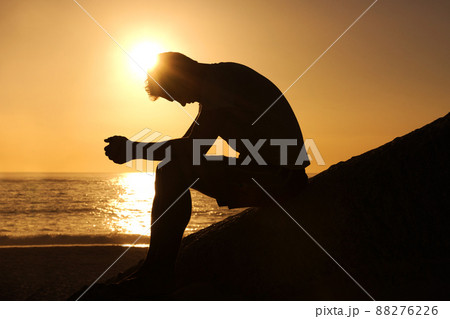 Contemplating the meaning of life. Silhouette of a man sitting by the ocean and contemplating while looking downwards. 88276226
