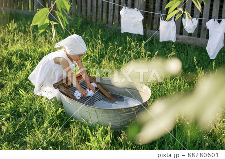Sweet girl in a kerchief and a dress launder snow-white linen by hand on an old washing board. Wet underwear for baby hanging on a clothesline at the village fence. Bleaching clothes. Sweet girl in a kerchief and a dress launder snow-white linen by hand on an old washing board. Wet underwear for baby hanging on a clothesline at the village fence. Bleaching clothes. 88280601