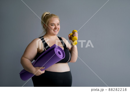 Fat Blonde woman holding pilates mat and yellow dumbbells. Fitness classes and a healthy lifestyle. Obese full-figured lady in black sportswear looks away copy space. Happy model body positive Fat Blonde woman holding pilates mat and yellow dumbbells. Fitness classes and a healthy lifestyle. Obese full-figured lady in black sportswear looks away copy space. Happy model body positive 88282218