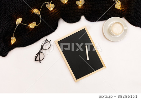 A cup of coffee, a small black wooden board and a white chalk with new years goals. Scarf and Christmas lights on white background. 88286151