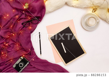 A cup of coffee, a small black wooden board and a white chalk with new years goals. Scarf and Christmas lights on white background. 88286153