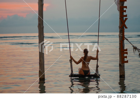 Tanned woman riding on a swing standing in the water. Sunset and sea in the background. Close up. Relax and vacation concept Tanned woman riding on a swing standing in the water. Sunset and sea in the background. Close up. Relax and vacation concept 88287130