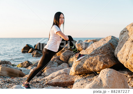 The concept of sport. A young brunette woman is engaged in stretching on the coastal rocks. In the background, the sea and the shore. Copy space The concept of sport. A young brunette woman is engaged in stretching on the coastal rocks. In the background, the sea and the shore. Copy space 88287227