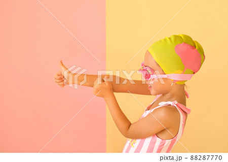 Cheerful positive girl in a trendy striped swimsuit, swimming glasses and a pool cap showing success gesture, thumb up with hand , saying well done. Pink-yellow studio background. Super cool swimming 88287770