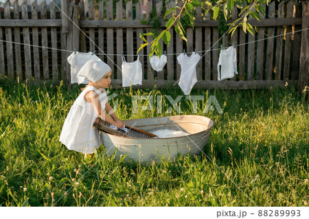 The child merrily washes his clothes in the basin. Splashes of water and foam from washing clothes. A girl in a white dress hangs out wet underwear to dry. Washing in an old basin. 88289993