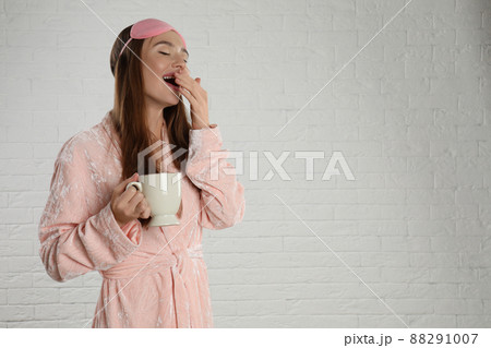 Beautiful young woman in bathrobe and eye sleeping mask yawning near white brick wall. Space for text Beautiful young woman in bathrobe and eye sleeping mask yawning near white brick wall. Space for text 88291007