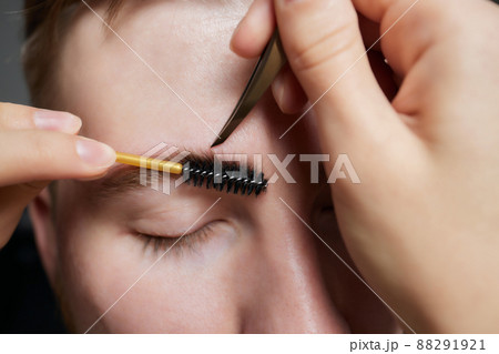 Professional barber doing threading procedure and correcting shape of eyebrows to young male client with tweezer in barber shop. Barber at work. Brow care concept. Plucking brows. Professional barber doing threading procedure and correcting shape of eyebrows to young male client with tweezer in barber shop. Barber at work. Brow care concept. Plucking brows. 88291921