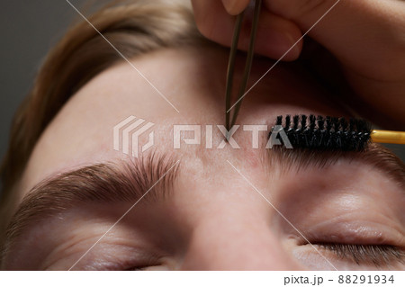 Professional barber doing threading procedure and correcting shape of eyebrows to young male client with tweezer in barber shop. Barber at work. Brow care concept. Plucking brows. 88291934