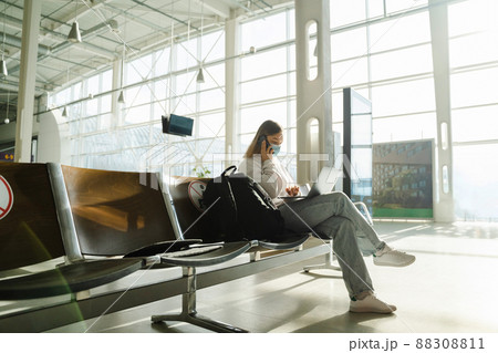 Female traveler in mask sits on chair in airport lobby with a social distance sign, talking on the phone and working on her laptop	 88308811