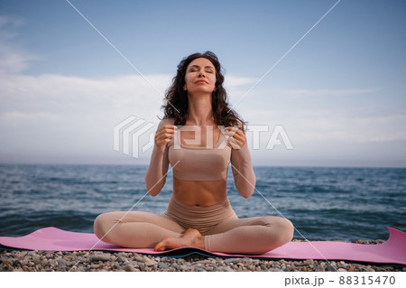 Well looking middle aged woman with black hair, fitness instructor in leggings and tops doing stretching and pilates on yoga mat near the sea. Female fitness yoga routine concept. Healthy lifestyle. Well looking middle aged woman with black hair, fitness instructor in leggings and tops doing stretching and pilates on yoga mat near the sea. Female fitness yoga routine concept. Healthy lifestyle. 88315470