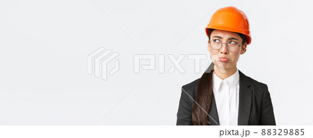 Close-up of gloomy upset asian female engineer in safety helmet and business suit complaining, looking disappointed, whining with sulking face, unwilling work on weekends, looking left bothered Close-up of gloomy upset asian female engineer in safety helmet and business suit complaining, looking disappointed, whining with sulking face, unwilling work on weekends, looking left bothered 88329885