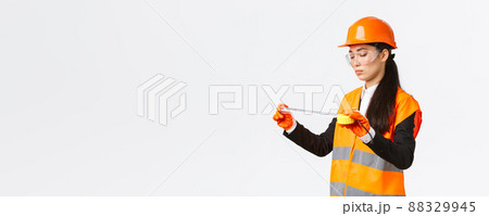 Serious asian female construction engineer, technician inspect layout, measuring something, looking at tape measure with focused face, standing over white background in safety uniform Serious asian female construction engineer, technician inspect layout, measuring something, looking at tape measure with focused face, standing over white background in safety uniform 88329945