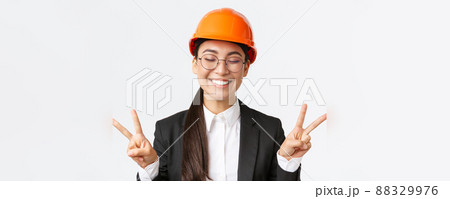 Close-up of cheerful successful female asian engineer, construction architect in safety helmet and business suit showing peace signs and smiling kawaii, standing white background 88329976