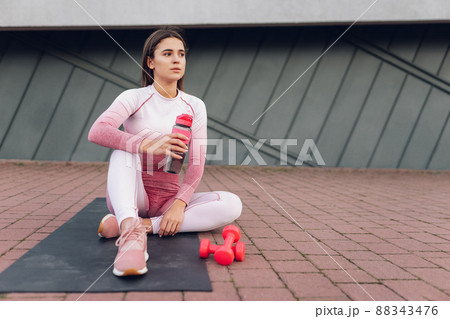 Fitness girl sitting with a bottle of water on the mat. Female athlete resting after training Fitness girl sitting with a bottle of water on the mat. Female athlete resting after training 88343476