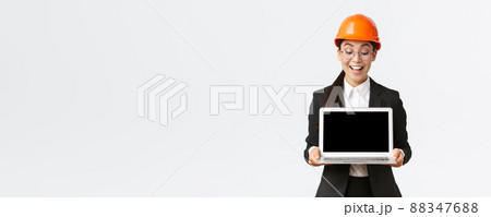 Impressed happy asian female chief engineer showing great results of enterprise to investors, standing in safety helmet and business suit, holding laptop and look pleased at computer screen 88347688