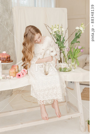 The girl is sitting at the Easter table with cakes, spring flowers and quail eggs and holding a white kid in her arms. Happy Easter celebration The girl is sitting at the Easter table with cakes, spring flowers and quail eggs and holding a white kid in her arms. Happy Easter celebration 88348139