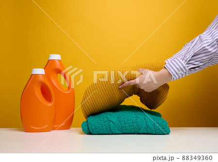 washed folded clothes and plastic orange large bottles with liquid detergent stand on a white table, yellow background. Routine homework washed folded clothes and plastic orange large bottles with liquid detergent stand on a white table, yellow background. Routine homework 88349360