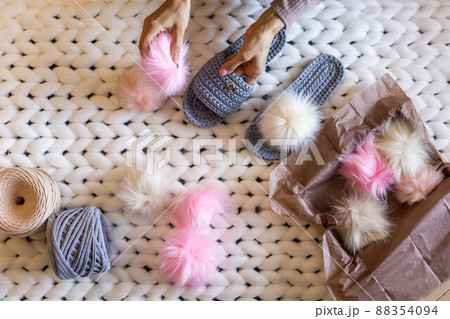 Woman hands connected fluffy fur pom pom to handmade ribbon yarn knitted slippers before sending Woman hands connected fluffy fur pom pom to handmade ribbon yarn knitted slippers before sending 88354094