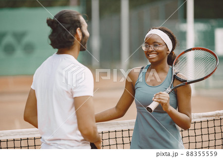 Indian man and black american woman standing on a tennis court. Woman wearing grey sport dress and man white t-shirt. Female and male tennis players talking. 88355559