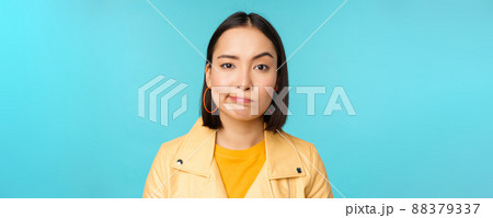Close up of skeptical asian girl looking disappointed, staring with doubt or disbelief, grimacing at camera, standing over blue background Close up of skeptical asian girl looking disappointed, staring with doubt or disbelief, grimacing at camera, standing over blue background 88379337