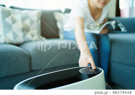 Happy Asian young woman turning on high efficiency air purifier while staying and reading a book in the living room. Woman using air purifier in her house. Happy Asian young woman turning on high efficiency air purifier while staying and reading a book in the living room. Woman using air purifier in her house. 88379438
