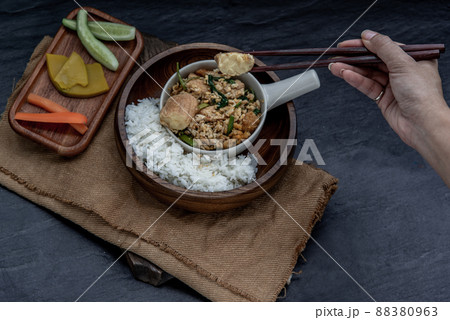 The hand is using chopsticks to pick up  Stir fried egg tofu with spring onion with Herbs vegetables served with steamed rice in wooden bowl. 88380963
