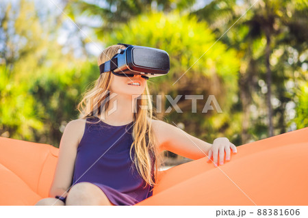 Summer lifestyle portrait of pretty girl sitting on the orange inflatable sofa and uses virtual reality headset on the beach of tropical island. Relaxing and enjoying life on air bed 88381606