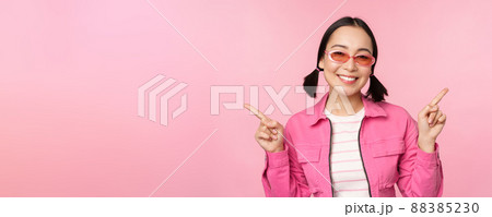 Choice. Stylish korean girl, asian female model points fingers sideways, shows two variants, product advertisement, demonstrating items, standing over pink background Choice. Stylish korean girl, asian female model points fingers sideways, shows two variants, product advertisement, demonstrating items, standing over pink background 88385230