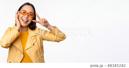 Portrait of stylish asian modern girl, wearing sunglasses and yellow jacket, showing peace, v-sign gesture, standing over white background, happy smiling face Portrait of stylish asian modern girl, wearing sunglasses and yellow jacket, showing peace, v-sign gesture, standing over white background, happy smiling face 88385282
