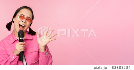 Happy beautiful asian girl singing with mic, using microphone, enjoying karaoke, posing against pink studio background Happy beautiful asian girl singing with mic, using microphone, enjoying karaoke, posing against pink studio background 88385284