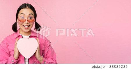 Beautiful asian girl smiling happy, showing heart gift box and looking excited at camera, standing over pink romantic background Beautiful asian girl smiling happy, showing heart gift box and looking excited at camera, standing over pink romantic background 88385285