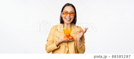 Stylish happy asian girl in sunglasses holding tangerines and smiling, posing against white background Stylish happy asian girl in sunglasses holding tangerines and smiling, posing against white background 88385287