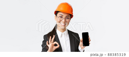 Satisfied young female asian engineer, architect in suit and safety helmet showing mobile phone screen and make okay gesture, approve, recommend application, white background Satisfied young female asian engineer, architect in suit and safety helmet showing mobile phone screen and make okay gesture, approve, recommend application, white background 88385319
