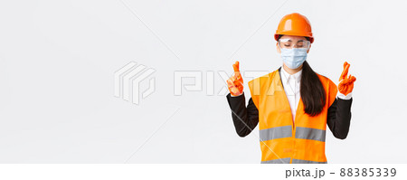 Covid-19 safety protocol at enterpise, construction and preventing virus concept. Hopeful asian female engineer in helmet, face mask praying, close eyes and cross fingers good luck, white background Covid-19 safety protocol at enterpise, construction and preventing virus concept. Hopeful asian female engineer in helmet, face mask praying, close eyes and cross fingers good luck, white background 88385339