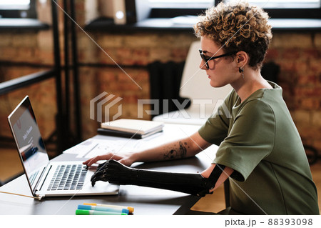 Young programmer with prosthetic arm working with software on laptop at her workplace at office 88393098
