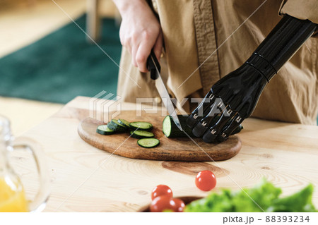 Close-up of woman with prosthetic arm cutting fresh cucumber with knife on the board at kitchen table 88393234