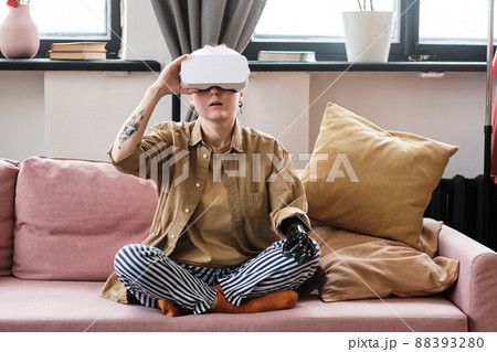 Young woman with arm prosthesis sitting on sofa in living room and using virtual reality headset Young woman with arm prosthesis sitting on sofa in living room and using virtual reality headset 88393280