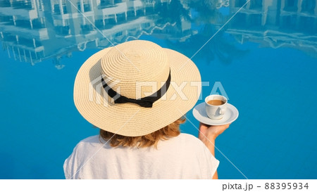 female in a straw hat holds a cup of coffee in her hand over a blue swimming pool concept of good female in a straw hat holds a cup of coffee in her hand over a blue swimming pool concept of good 88395934