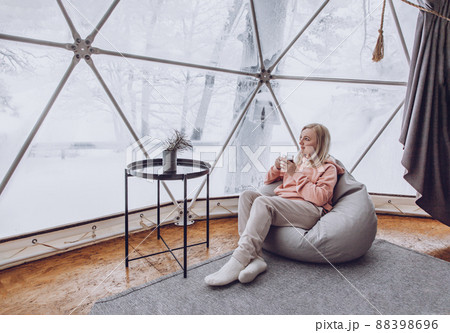 A woman is sitting in a geo dome glamping tent in a bag chair and looking out at the winter snow-covered nature and drinks coffee. A woman is sitting in a geo dome glamping tent in a bag chair and looking out at the winter snow-covered nature and drinks coffee. 88398696