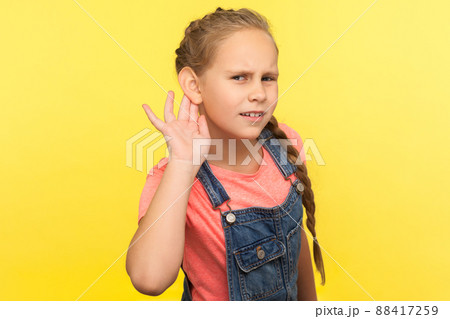 What. I can't hear. Portrait of curious attentive little girl in denim overalls holding hand near ear and listening carefully intently to what you say. indoor studio shot isolated on yellow background What. I can't hear. Portrait of curious attentive little girl in denim overalls holding hand near ear and listening carefully intently to what you say. indoor studio shot isolated on yellow background 88417259