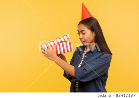 Portrait of curious happy brunette girl in denim shirt and funny cone hat opening birthday gift, looking inside with interest, unpacking present on party celebration. indoor studio shoot isolated 88417337