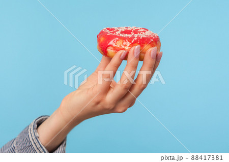 Closeup of female hand holding delicious glazed red donut with sprinkles, showing tasty doughnut, sweet confectionery high in glucose and calories. indoor studio shot isolated on blue background 88417381