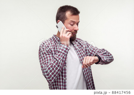Portrait of punctual bearded man in plaid shirt talking on phone and checking time on wristwatch, dissatisfied with delay, deadline, mobile communication. indoor studio shot isolated, white background 88417456