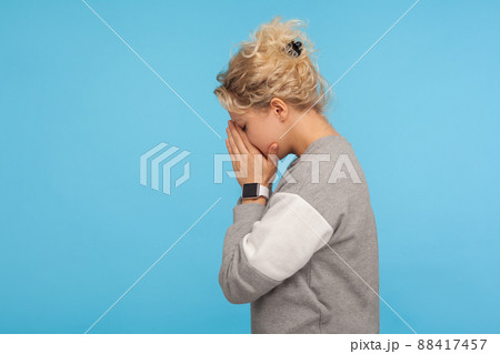 Side view of depressed woman with short curly hair in casual sweatshirt hiding face down in hands and crying, hard going through loss or defeat, feeling sorrow. studio shot isolated on blue background 88417457