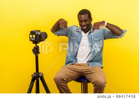Here and right now, online business training. Portrait of happy friendly man in denim shirt smiling at his professional dslr camera and pointing down, asking subscribe now. indoor studio shot isolated 88417458