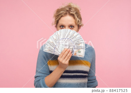 Portrait of positive woman with curly hair in sweater holding dollar banknotes, peeking out of money while hiding face, looking surprised by big profit. indoor studio shot isolated on pink background 88417525