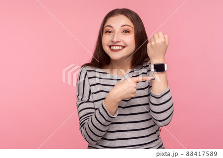Hurry up. Portrait of positive woman in striped sweatshirt pointing smartwatch on her wrist and looking at camera with toothy smile, worried about late time. studio shot isolated on pink background 88417589