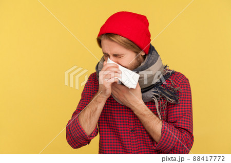 Sick guy in hat and warm scarf coughing sneezing napkin, feeling unwell with runny nose, sore throat, suffering allergy or seasonal influenza symptoms. indoor studio shot isolated on yellow background Sick guy in hat and warm scarf coughing sneezing napkin, feeling unwell with runny nose, sore throat, suffering allergy or seasonal influenza symptoms. indoor studio shot isolated on yellow background 88417772