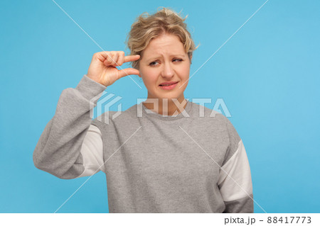 Need some more. Displeased disappointed woman with short curly hair in sweatshirt showing minimum sign with fingers, small size, little bit gesture. indoor studio shot isolated on blue background 88417773