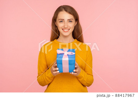Portrait of beautiful cheerful red hair girl in sweater standing holding wrapped gift box and looking at camera joyfully with smile, festive mood. indoor studio shot isolated on pink background Portrait of beautiful cheerful red hair girl in sweater standing holding wrapped gift box and looking at camera joyfully with smile, festive mood. indoor studio shot isolated on pink background 88417967
